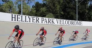 An image of track cyclists riding in a line on a velodrome. They are wearing red and black. A sign above them says "Hellyer Park Velodrome".