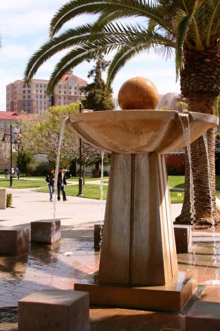 Image of a fountain with a large column with a bowl of water on top and a ball on top of the bowl. Water is spilling out of the bowl. There is a palm tree in the background. 