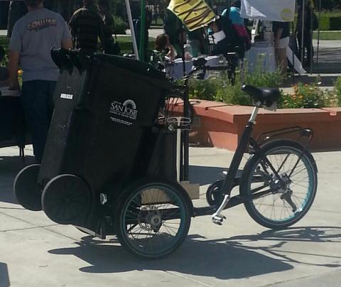 An image of a black three-wheeler bicycle with a garbage bin on the front of it. 