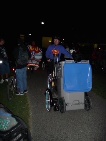 An image of a person wearing a superman shirt riding a three-wheeler bike with a garbage or recycling bin in the front of it. 