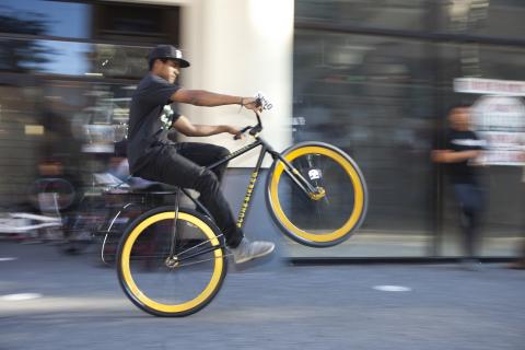 An image of a person doing a wheelie on a fixed gear bicycle. The person is wearing all black with a black baseball cap, and the bicycle has a black frame with yellow tire rims.