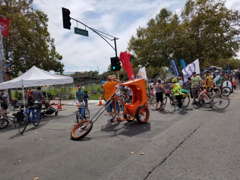 An image of a group of people with bicycles. In the foreground is a person wearing orange with an orange super low rider three-wheeler bicycle, with an extended fork and front wheel, and a cockpit with a covered chariot.