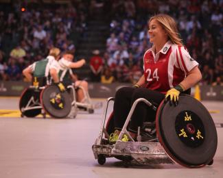 A white person with long blond hair is smiling while playing wheelchair basketball on a wooden court. The person is wearing black pants and a red jersey with the number 24 on it.  