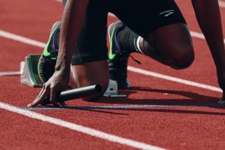 A runner is in the start position before a race on a red track. They are Black, and they are holding baton and wearing black shorts, black, purple and green Nikes. Their head is not visible in the photo. 